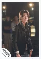 山下智久/上半身・衣装黒・スカーフ・両手下げ・体右向き・カメラ目線・後ろに人・背景黒・ロゴ「YAMASHITA TOMOHISA」/公式生写真