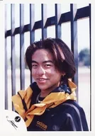 V6 / Tsuyoshi Morita / Bust up / Costume black / yellow / Both hands down / Teeth show / Camera line of sight / Body facing left / Outdoor / Back fence / Official Official photo